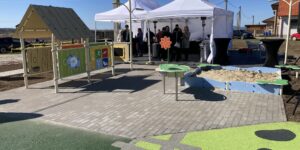 Outdoor event with white canopy tents, people socializing near a children's play area against a bright blue sky.
