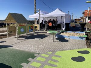 Outdoor event with white canopy tents, people socializing near a children's play area against a bright blue sky.