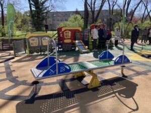 Colorful train-themed playground with slides and climbing frames; adults stand nearby under trees in the background.
